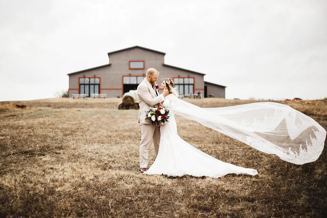 Bride and groom embracing in a field with bridal veil flowing, barn structure visible in background