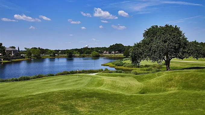 Golf course with manicured fairways, water hazard, and large tree under blue sky with white clouds