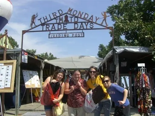 Visitors walking through outdoor vendor stalls under a metal arch sign reading "Third Monday Trade Days" with trees and tents visible
