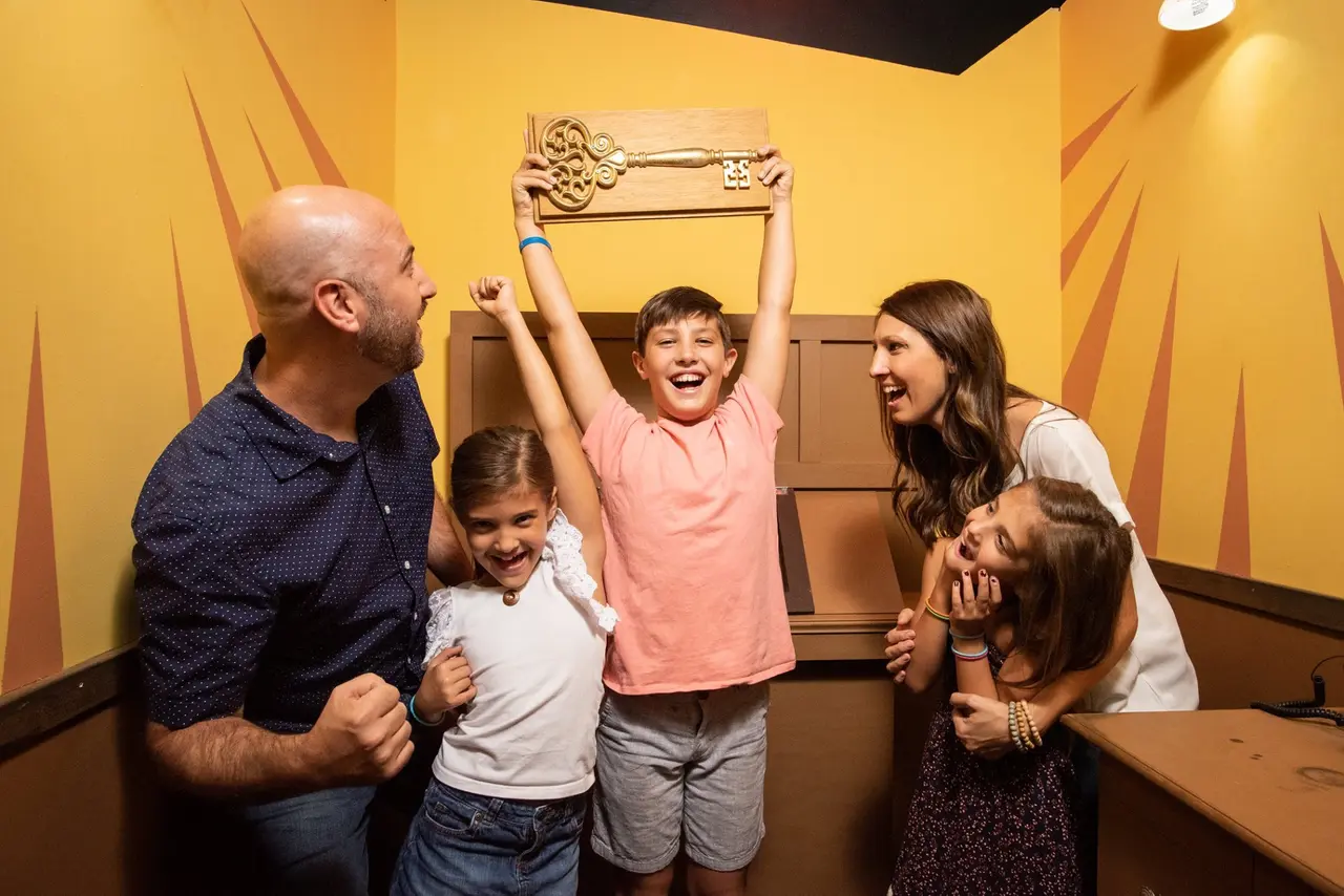 Family of five celebrating with raised arms in bright yellow-walled escape room with decorative door frame