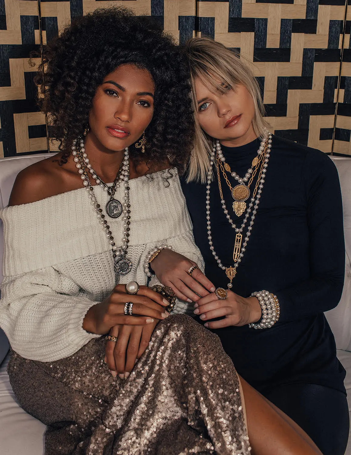 Two women wearing layered pearl and metal chain necklaces and multiple rings, posing together against a geometric patterned backdrop