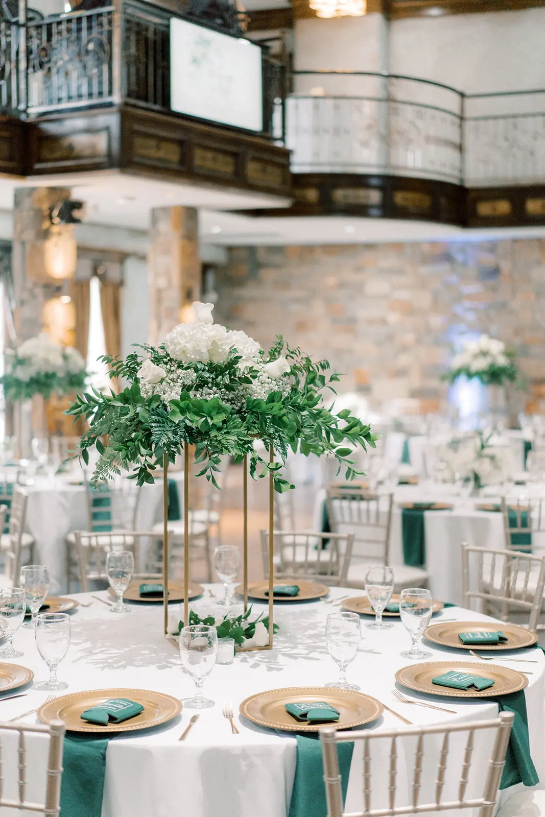 Elegant banquet table with white linens, gold chargers, teal napkins, and tall floral centerpiece with white flowers and greenery in an ornate brick venue