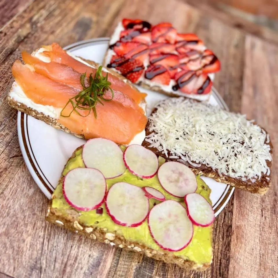 Open-faced sandwiches with smoked salmon, radish slices on avocado spread, and shredded cheese on rye bread on wooden surface
