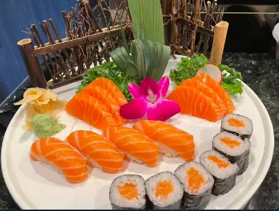 Sashimi and sushi rolls arranged on a white plate with fresh orange slices, pink radish flower, and green garnish