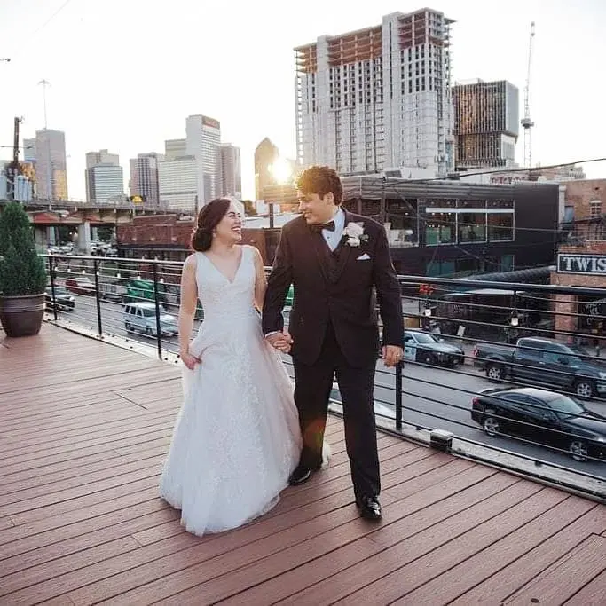 Bride and groom holding hands on rooftop deck with downtown city skyline at sunset in background