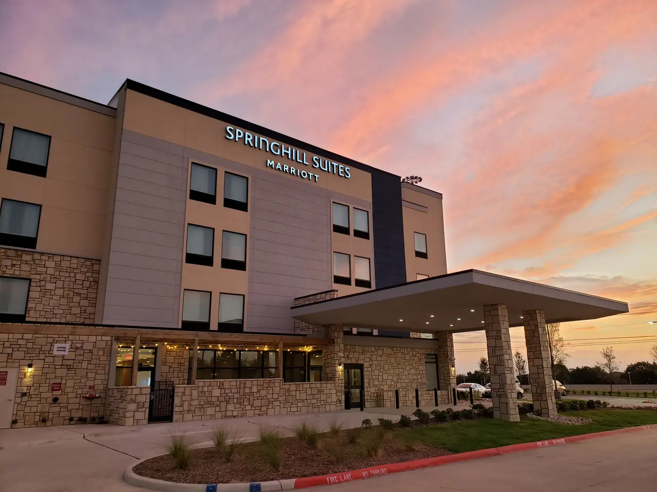Modern hotel building with brick and siding exterior, illuminated entrance canopy, and pink-orange sunset sky at dusk