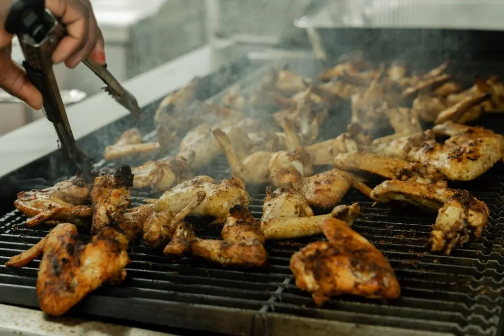 Hand holding tongs over grill with seasoned chicken pieces cooking over flame, smoke rising