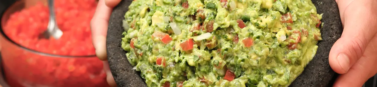 Hand holding dark bowl of guacamole with diced tomatoes and cilantro, red salsa visible beside it