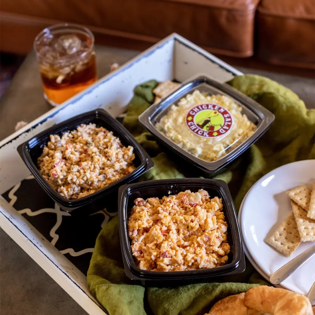 Three black containers of chicken salad varieties with crackers and a beverage on a white tray