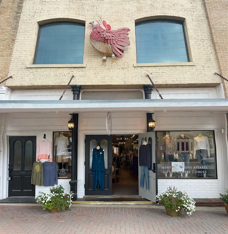 Brick storefront with large pink rooster sculpture above two blue-framed windows, black doors, clothing displays, and potted white flowers
