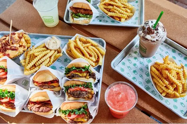 Multiple burgers and waffle fries on white plates, with cold beverages on a wooden table