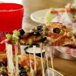 Slice of pizza with melted cheese and toppings being pulled from whole pizza, red cup and plate in background