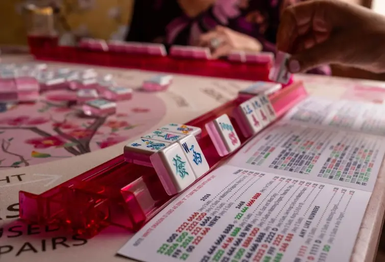 Players arranging mahjong tiles on racks during a game, with score sheets and instruction materials visible on the table