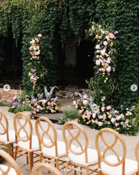 Ivy-covered brick archway decorated with peach and white roses, wooden chairs in foreground