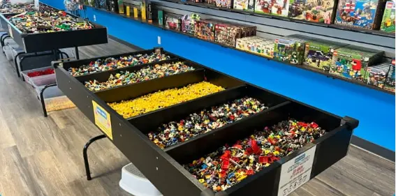 Large black bins filled with colorful LEGO bricks and minifigures displayed in front of blue shelving with boxed sets
