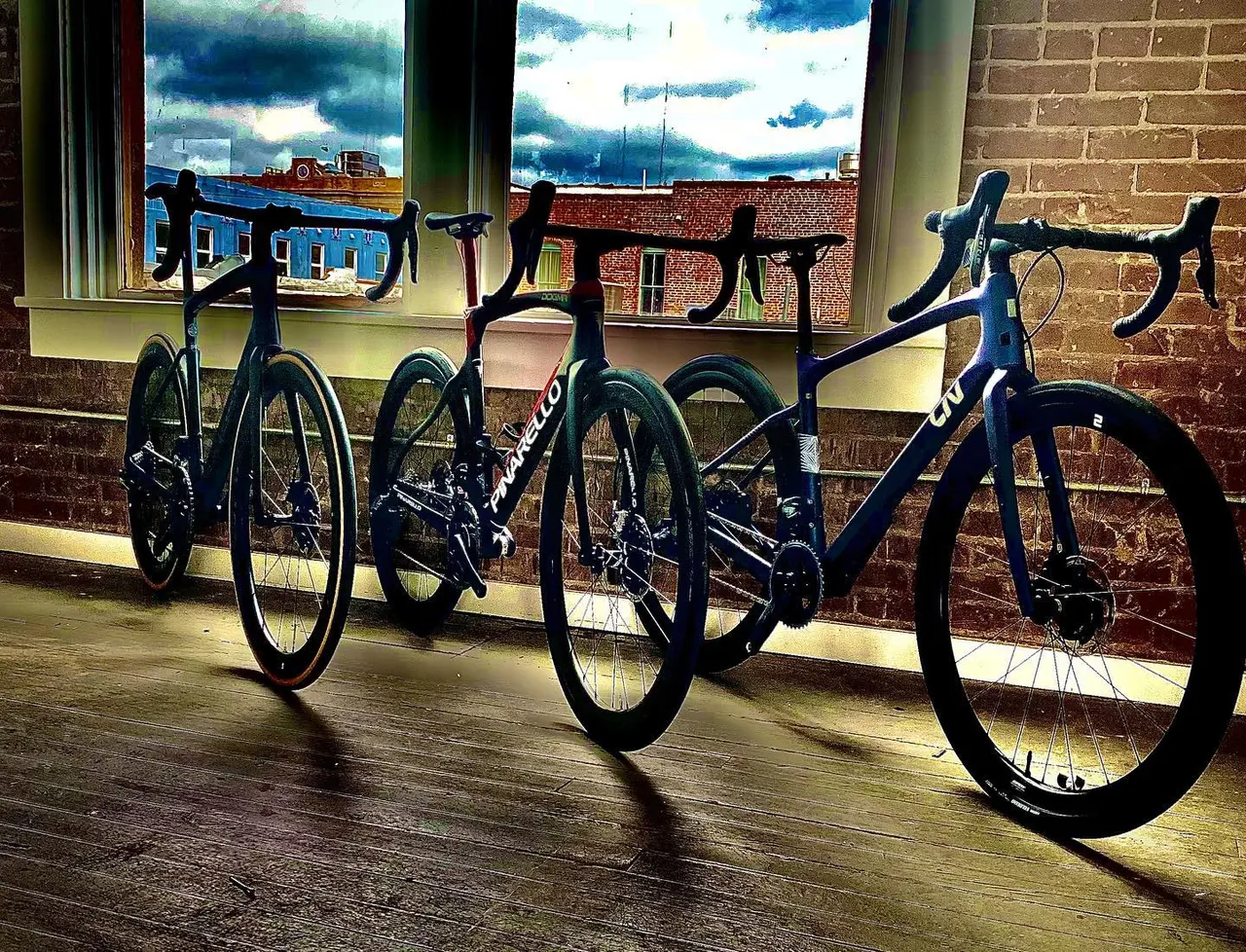 Three road bikes lined up indoors with large windows overlooking urban rooftops and cloudy sky