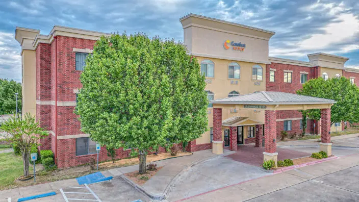 Modern hotel building with red brick and tan facade, green trees, covered entrance, and parking area under blue sky