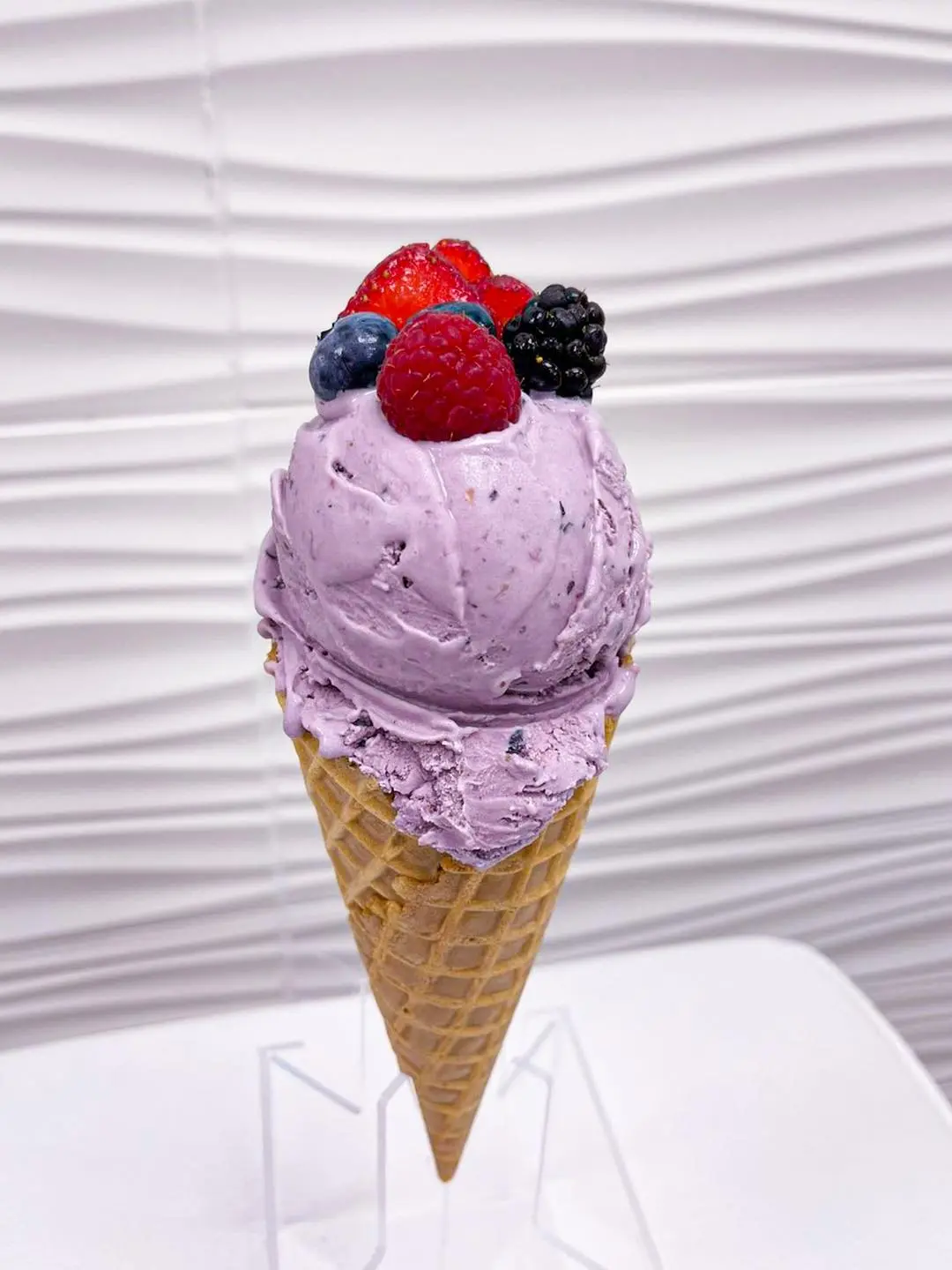 Purple ice cream cone topped with fresh raspberries, blueberries and blackberries against white textured background