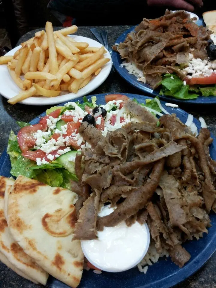 Plates of Greek food including sliced meat with pita bread, salad with feta cheese, white sauce, and french fries