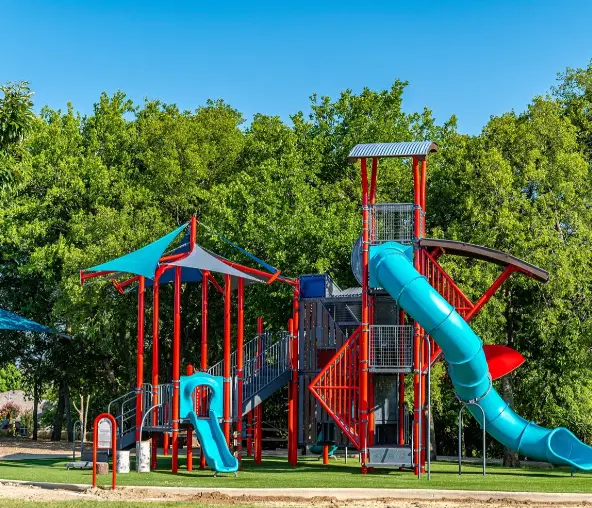 Bright red and blue playground structure with multiple slides and climbing features surrounded by tall green trees under clear sky