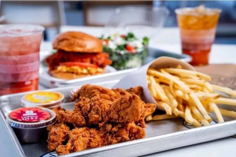 Fried chicken pieces and fries on a white plate with sauce cups, alongside a chicken sandwich and salad at an outdoor dining setting
