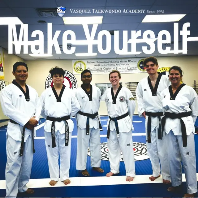 Group of six taekwondo students in white uniforms and black belts standing together in a studio