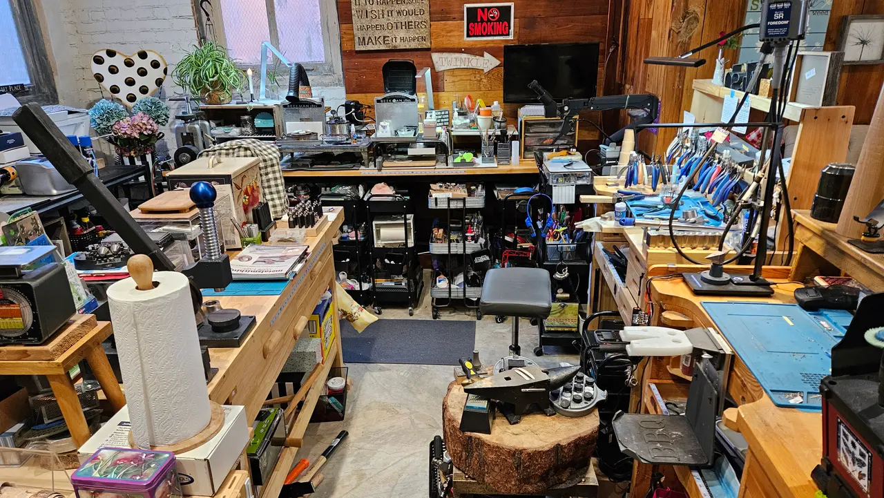 Cluttered jewelry studio interior with wooden workbenches, shelving, tools, materials, and a tree stump base visible throughout the space