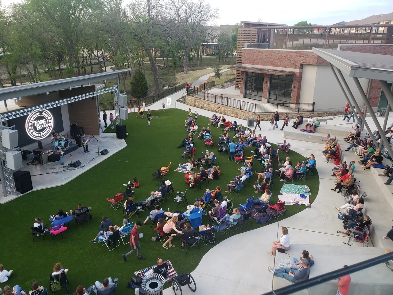 Aerial view of outdoor courtyard with green lawn, crowds of people seated at tables, stage with performance setup, and brick buildings surrounding the space