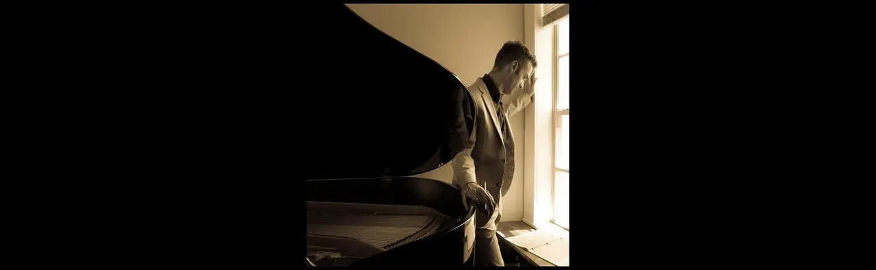Man looking out of a window with a piano at his back. Photo is in black and white and is moody