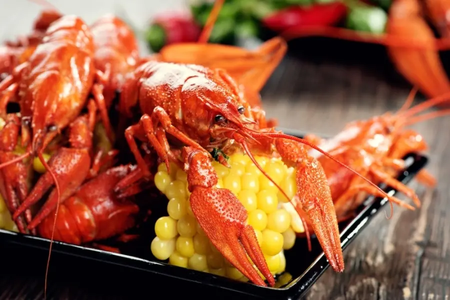 Boiled crawfish with yellow corn on a black plate, with additional seafood and vegetables blurred in background