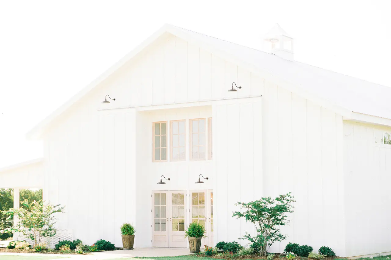 Modern white barn-style building with double doors, large windows, and landscaped entrance featuring potted plants and shrubs