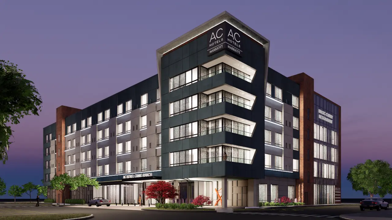Modern multi-story hotel building with mixed brick and dark facade, illuminated ground-floor retail spaces, and purple twilight sky behind