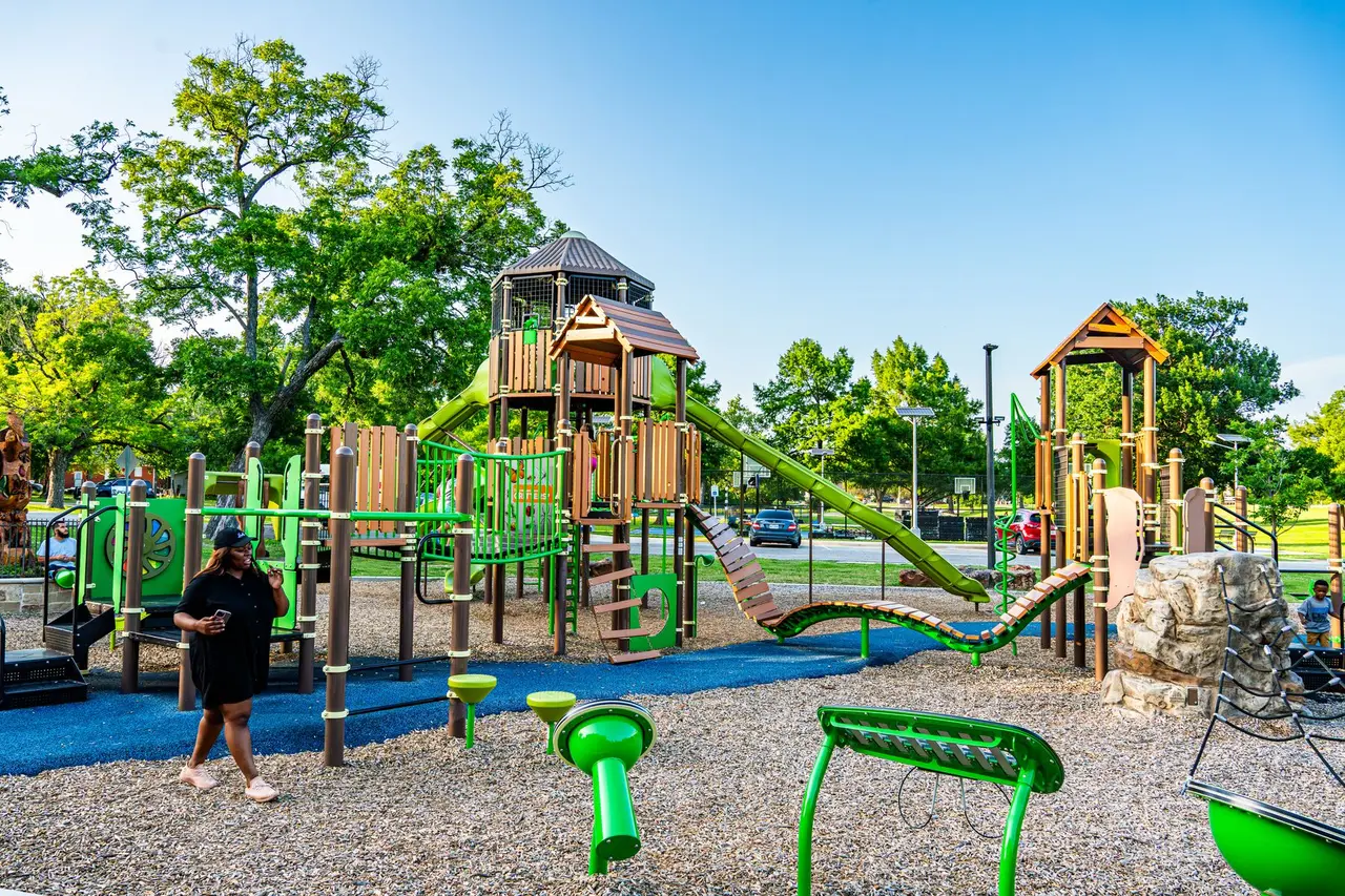 Colorful playground with green equipment and climbing structures, surrounded by trees and blue safety surfacing