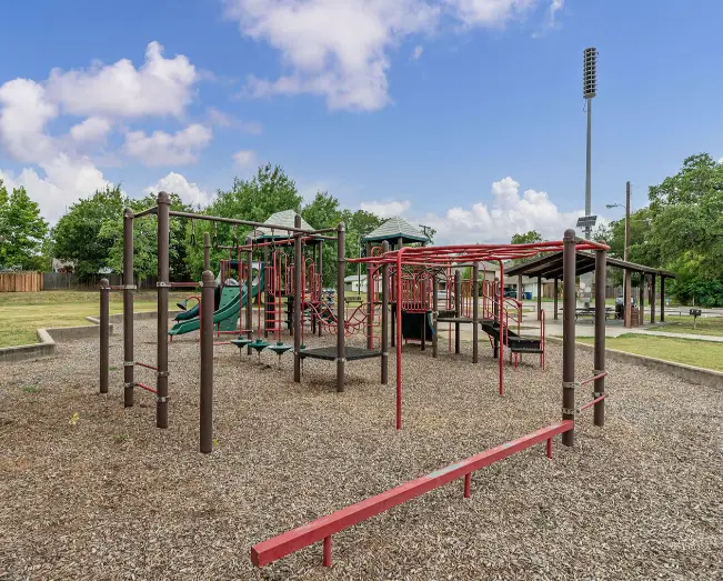 Large red and brown playground structure with multiple levels and climbing features on mulch surface, surrounded by wooden posts and benches, with green lawn