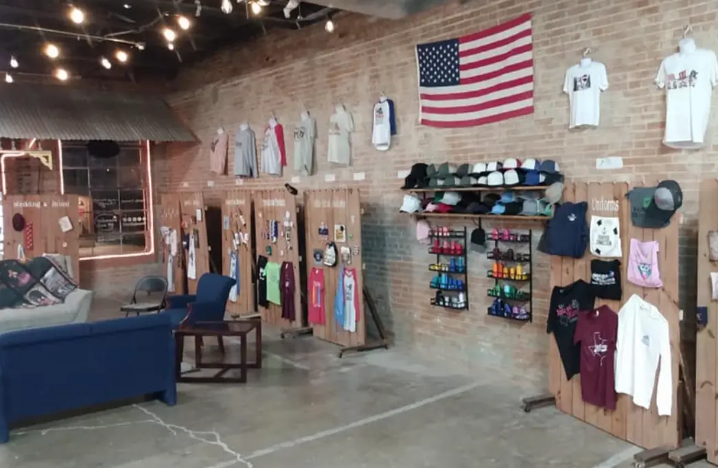 Retail shop interior with exposed brick wall displaying hanging t-shirts, apparel, and merchandise organized on wooden pegboards, with an American flag and