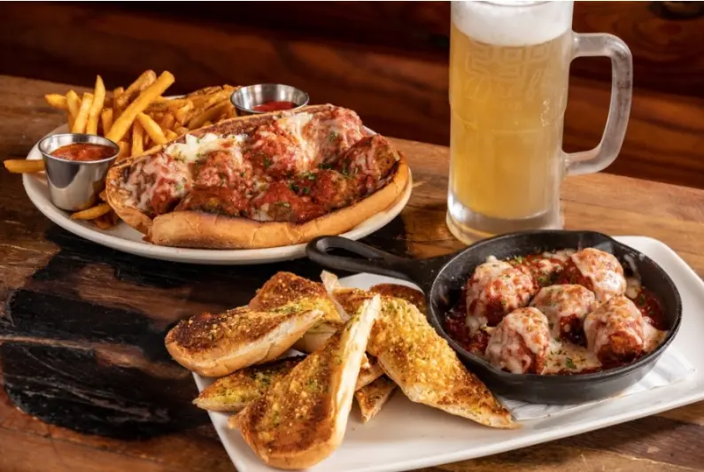 Fried chicken sandwich with fries, garlic bread, meatballs in skillet, and glass mug of beer on wooden table
