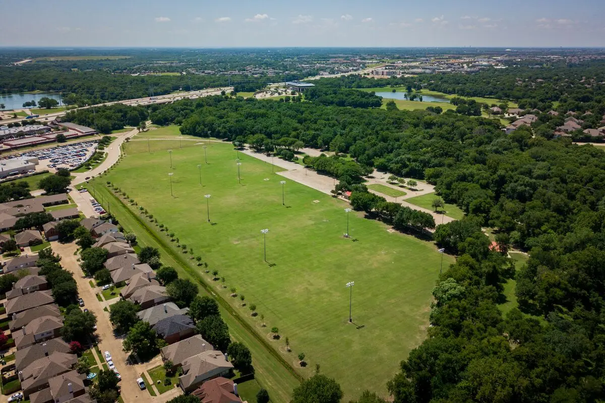 Aerial view of large green soccer fields with rows of trees and residential neighborhoods visible on the left and wooded areas on the right