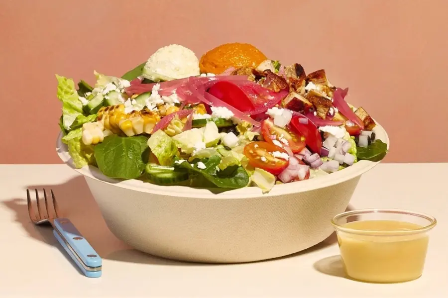 Large salad bowl with mixed greens, roasted vegetables, grains, feta cheese, and pickled red onions, served with dressing and fork