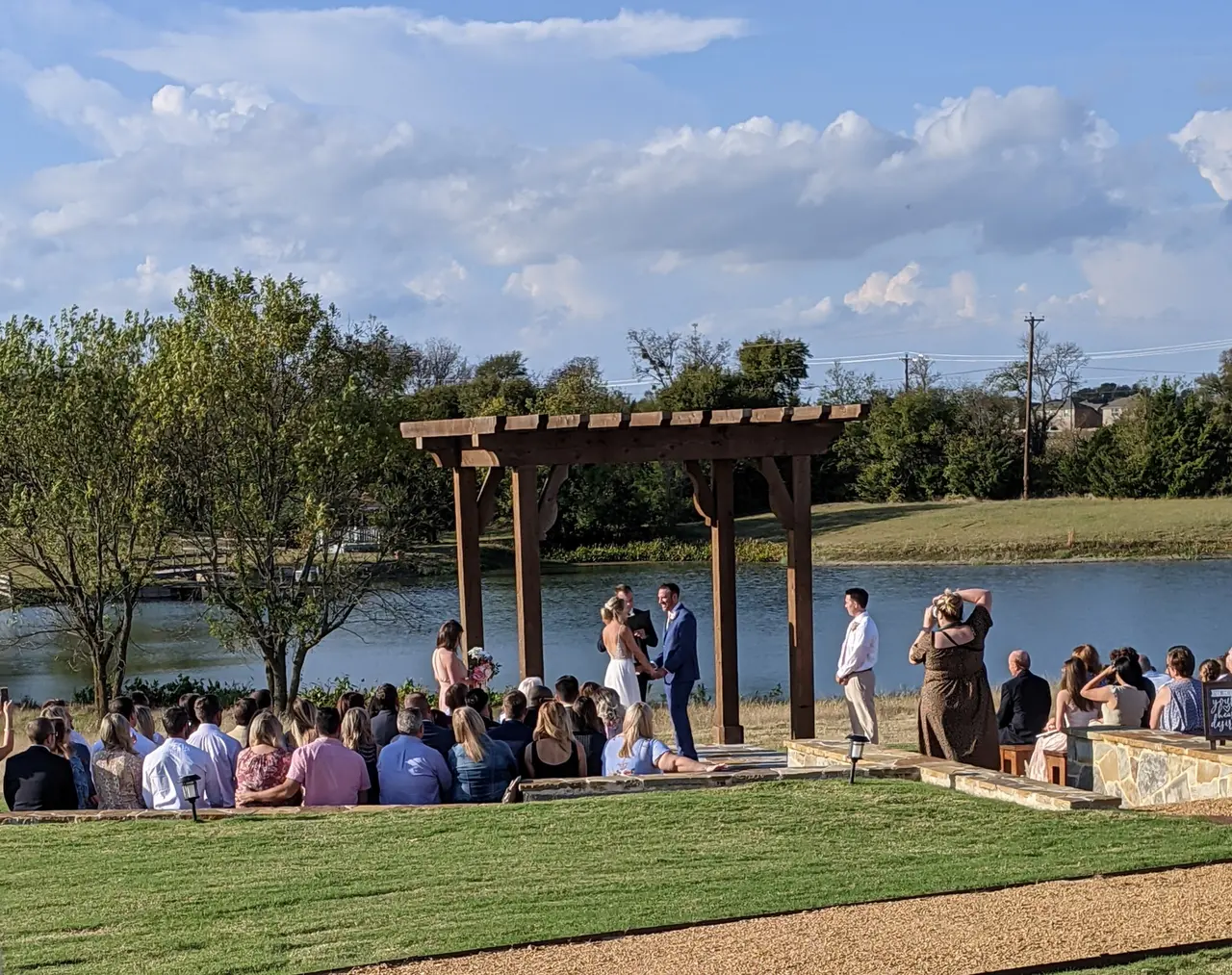 Outdoor wedding ceremony under wooden pergola beside a pond, with guests seated on lawn and couple exchanging vows
