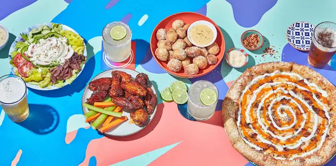 Colorful overhead view of pizza, fried chicken, appetizers, and beverages arranged on a bright blue and pink background