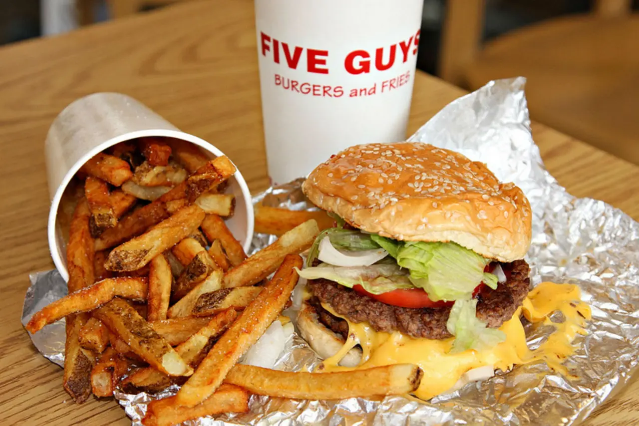 Cheeseburger with lettuce and tomato on sesame bun, cajun-style fries in paper cup, and branded cup on wooden table