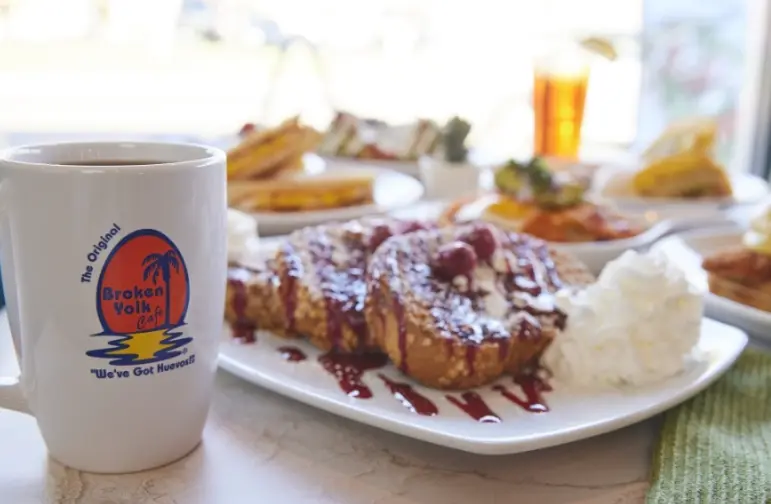 Coffee mug beside a plate of French toast with berries and whipped cream, with pastries and beverages blurred in background