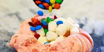 Hand holding pink ice cream topped with colorful candy pieces spilling from a container above