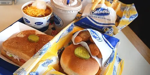 Culver's meal spread including a butter burger with pickles, cheese curds, frozen custard in branded cups, and fries in branded packaging on a table