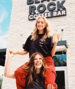 Two women posing in front of a white building with Black Rock Coffee Bar signage, one seated on the other's shoulders with arms raised