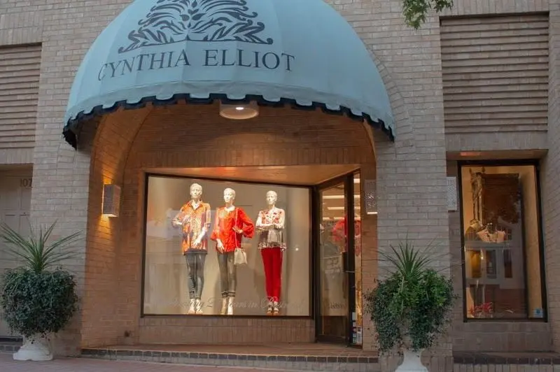 Storefront with blue awning displaying mannequins in colorful clothing in illuminated window, flanked by potted plants