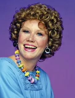 Woman with curled blonde hair wearing a light blue caftan and colorful beaded necklace against purple background