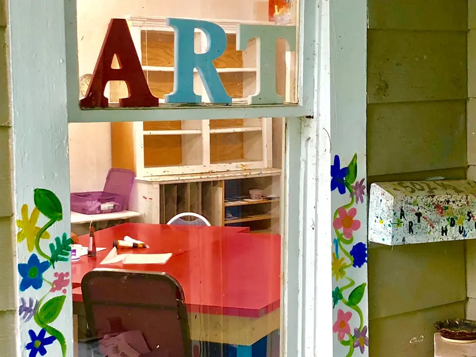 Colorful art studio interior with large blue and red letters spelling "ART" above shelving units and a desk with chair