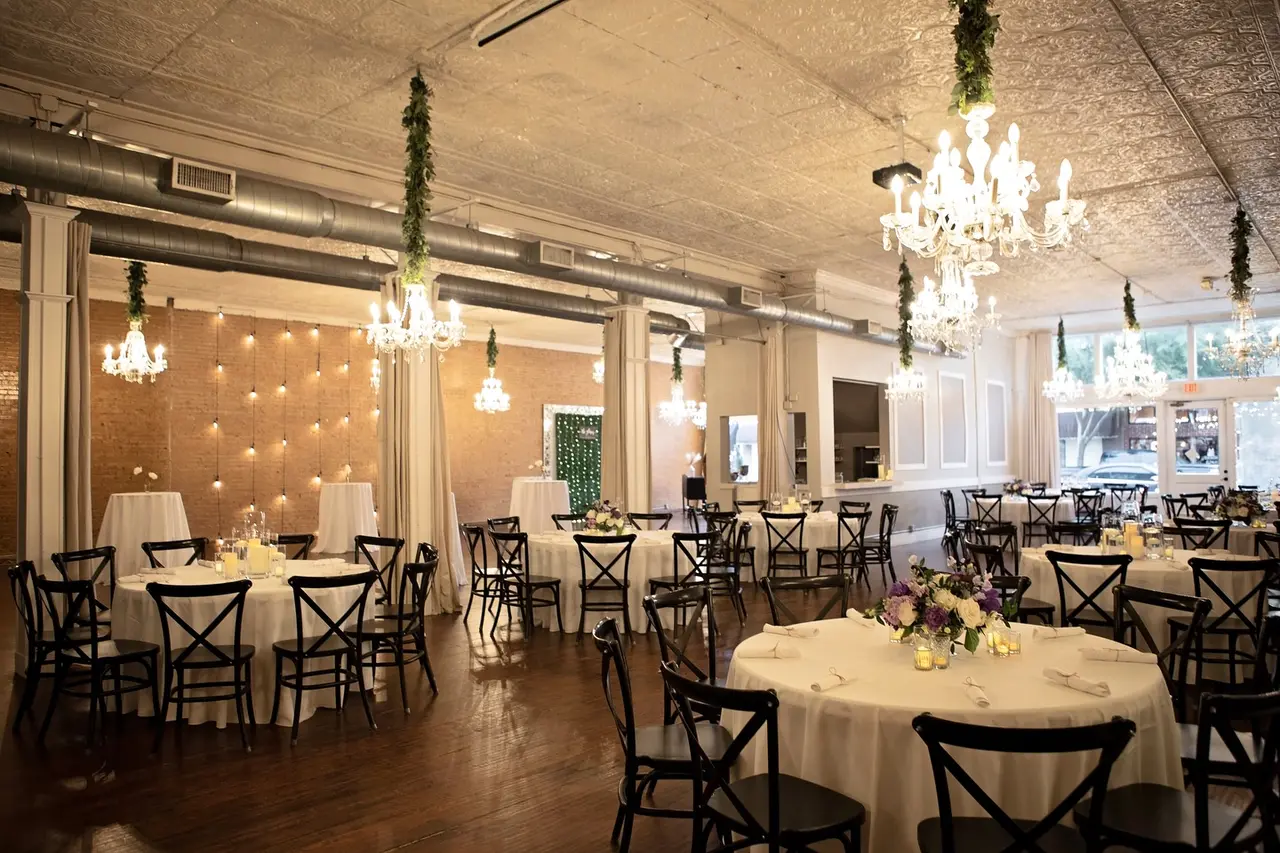 Elegant event space with round tables, black chairs, crystal chandeliers, and warm lighting against exposed brick walls