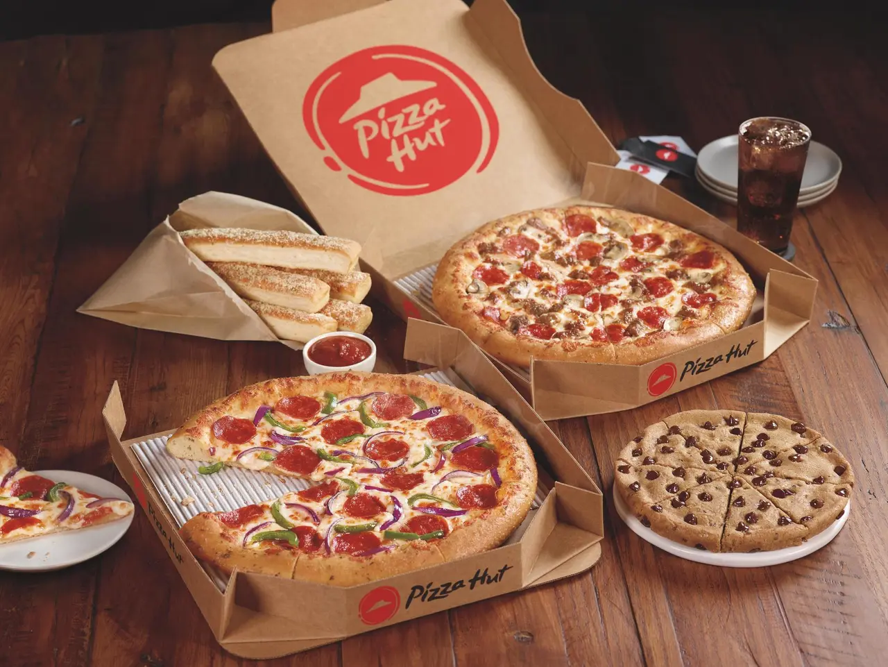 Two pepperoni pizzas in cardboard boxes, breadsticks with dipping sauce, a dessert pizza, and a glass of cola on a wooden table